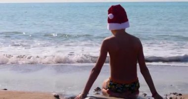 Sırtı büyük parlak Noel Baba şapkalı mutlu çocuk deniz kıyısında oturur ve kumla oynar, güneşe karşı.