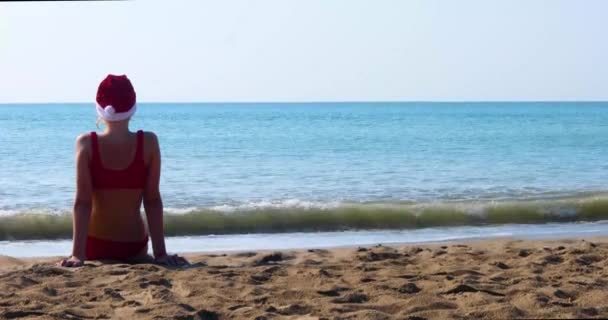身穿红色泳衣 头戴闪亮的桑塔爪帽的少女 背对着阳光坐在海滨 望着海浪 元旦假期 — 图库视频影像