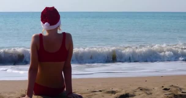 快乐的女孩 背对着阳光 戴着闪亮的圣诞老人帽 坐在海滨 元旦假期 — 图库视频影像