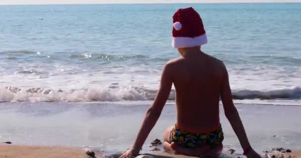 太陽に対して 海の海岸に座って砂と遊ぶ大きな光沢のあるサンタクラスの帽子で彼の背中を持つ幸せな少年 — ストック動画