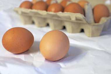 two eggs on a table near brown eggs in a cardboard container on a light background.