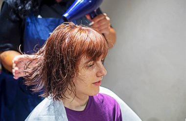 Kuaförün elleri bir saç kurutma makinesini tutuyor ve açık arkaplandaki bir kuaförde saç kesiminden sonra kızıl saçlı bir kızın ıslak saçını kurutuyor.
