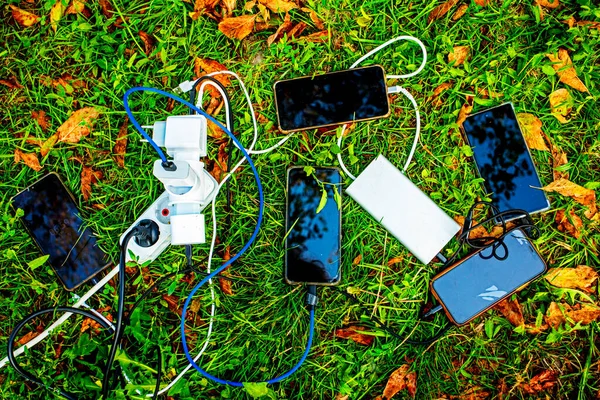 five smartphones charging from an outlet on the grass, flatlay. Family trip in nature, camping