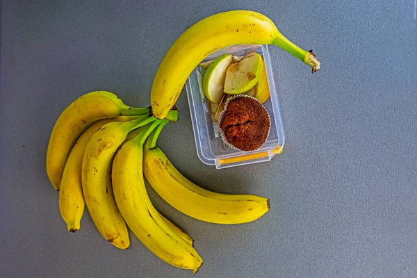 bunch of bananas on the kitchen table next to a container with apple slices and muffins and a school notebook with pens. school snack