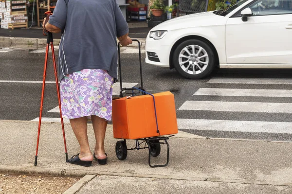 elderly woman with walking sticks and a cart for things stands on a crosswalk. Caring for the health of the older generation
