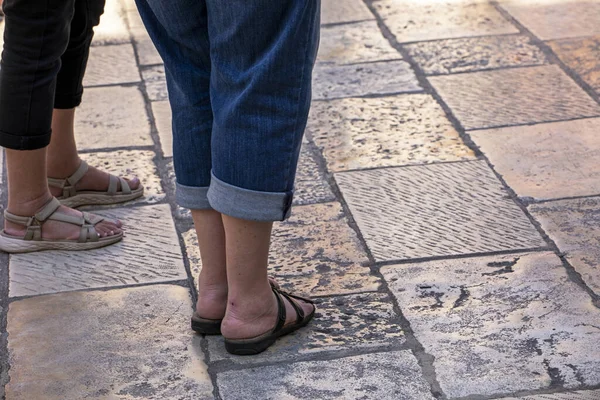 swollen feet of women in flip-flops stand on shiny masonry, tourists and travel