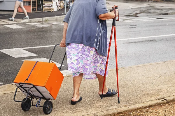 elderly woman with a stick is pushing a cart with a box. Caring for the Elderly