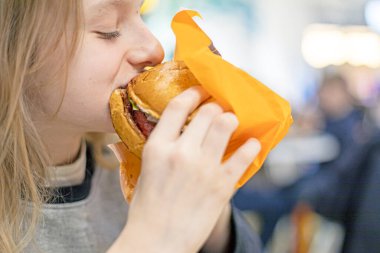 schoolgirl girl eats a vegetarian cheeseburger in gloves during recess. school snack. schoolchildren and teenagers meals clipart