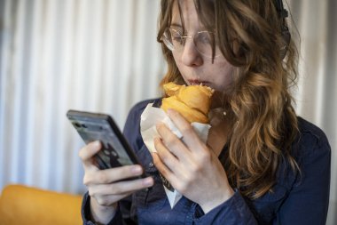 Genç kız kruvasan yer ve akıllı telefondan mesajlar okur. Modern teknolojiler. Sanal iletişim