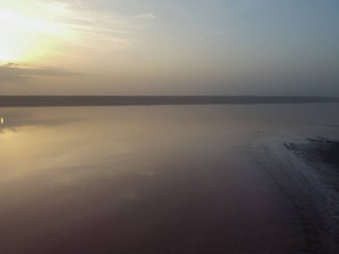 Fotoğraf, kumlu bir sahili olan kırmızı bir tuz gölünü akşam ışığında ufka uzanırken gösteriyor. Gölün koyu kırmızı renkleri çevresindeki toprak ve bitki örtüsüyle çok güzel tezat oluşturuyor., 