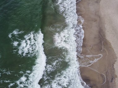 İnsansız hava aracı, kumlu bir sahile vuran dalgalarla denizin çarpıcı bir görüntüsünü yakaladı. Kristal berrak su ve beyaz köpük dalgalar büyüleyici bir şekil oluşturur.