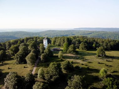 Ufuk manzaralı yeşil bir parkta beyaz bir kule. Kule ağaçlarla çevrili ve oraya giden bir yol var. Park bir tepede ve gökyüzü açık ve mavi. Alman kırsal kesimi