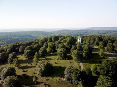 Ufuk manzaralı yeşil bir parkta beyaz bir kule. Kule ağaçlarla çevrili ve oraya giden bir yol var. Park bir tepede ve gökyüzü açık ve mavi. Alman kırsal kesimi