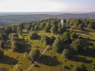 Ufuk manzaralı yeşil bir parkta beyaz bir kule. Kule ağaçlarla çevrili ve oraya giden bir yol var. Park bir tepede ve gökyüzü açık ve mavi. Alman kırsal kesimi