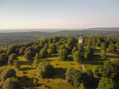Yeşil ormanlık alandaki beyaz bir kulenin havadan çekilmiş fotoğrafı. Kule yüksek ve dikdörtgen bir çatı ve tepesinde bir haç var. Kule ağaçlarla çevrili ve ona giden toprak bir yol var. Alpler.