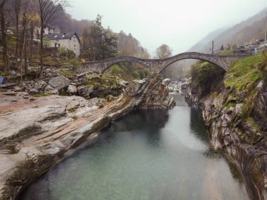 Dağlık bir bölgedeki bir nehrin üzerindeki taş bir köprünün fotoğrafı. Köprü yeşilliklerle kaplı ve aşağıdaki nehir kayalık bir geçitten akıyor. Arka plan sisli bir dağdan oluşuyor.