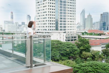 Yalnız Asyalı kadın, açık hava molası sırasında koruyucu maske takıyor ve şehrin arka planında çatıda rahatlıyor.