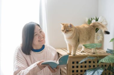 Asyalı kadınla kedi bakımı konsepti kitap okumak ve altın rengi İngiliz kedisiyle evde oynamak