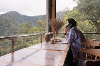 Dijital göçebe konsepti Asyalı serbest çalışan kadın dağ manzaralı kahve dükkanında çalışmak için akıllı telefon kullanıyor.