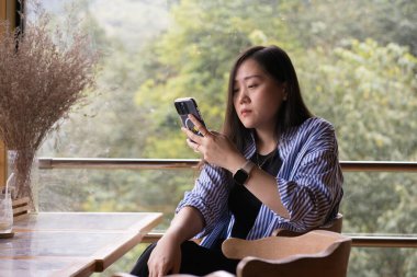 Dijital göçebe konsepti Asyalı serbest çalışan kadın dağ manzaralı kahve dükkanında çalışmak için akıllı telefon kullanıyor.