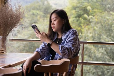 Dijital göçebe konsepti Asyalı serbest çalışan kadın dağ manzaralı kahve dükkanında çalışmak için akıllı telefon kullanıyor.