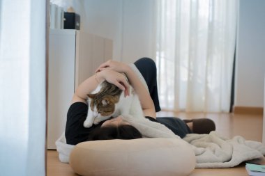 Asyalı kadınla sağlık ve refah kavramı uzanıp kitap okuyor ve kedisiyle oturma odasında oynuyor.