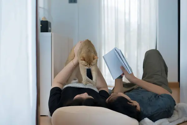 wellness and wellbeing concept with asian couple lie down and read book and play with their cat in livingroom