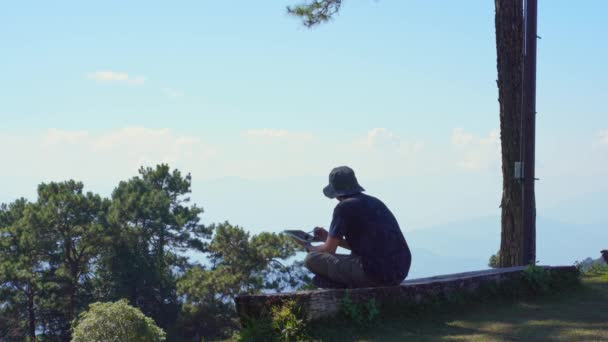 ソロフリーランサーの男性とデジタルノマドと旅行のコンセプトは木に座って 山の背景の層で屋外からタブレットワークを使用します — ストック動画