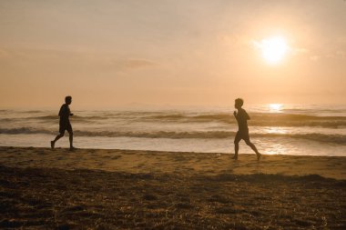 Siluet iki adam sahilde koşuyor, gün doğumu ve deniz altyapısı var.