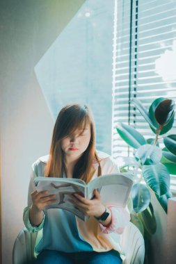 Genç ve güzel Asyalı kadın evinde rahat ve rahat bir şekilde kitap okurken keyif alır.