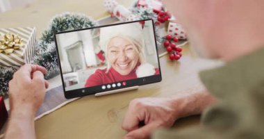 Kafkasyalı bir adam, noel süslemeleri ile beyaz bir kadınla tablet video görüşmesi yapıyor. Noel, kutlama, dijital kompozit video.