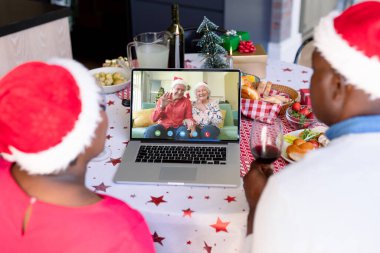 Kıdemli Afro-Amerikan çifti, Kafkasyalı bir çiftle Noel 'de görüntülü görüşme yapıyor. İletişim teknolojisi ve noel, dijital kompozit görüntü.