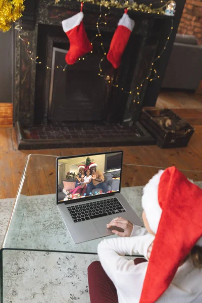 幸せなアフリカ系アメリカ人の家族とクリスマスのラップトップビデオ通話を持つサンタの帽子を持つ白人女性 クリスマス お祝いとデジタル複合画像 — ストック写真