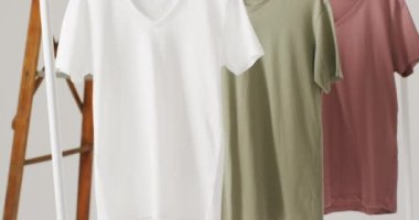 Beyaz arka planda asılı beyaz, bej ve gren tişörtlerin yakın çekim videosu. Giyim, moda, perakende, kumaş, doku ve malzeme konsepti.