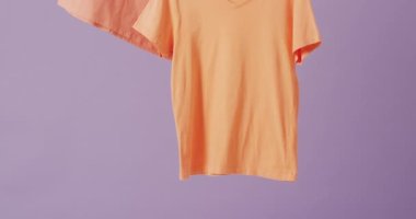 Mor arkaplanda asılı turuncu tişörtlerin yakın çekim videosu. Giyim, moda, perakende, kumaş, doku ve malzeme konsepti.