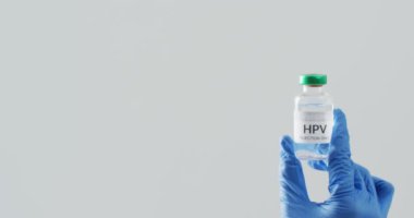 Elinde ameliyat eldiveni olan el kamerasının gri arka planda fotokopi alanı olan HPV aşı şişesi tutarken videosu. Tıp, sağlık hizmetleri, sağlık hizmetleri ve sağlık farkındalığı kavramı.