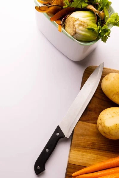 Vertical of knife and vegetables on chopping board with vegetable waste in kitchen composting bin. On white background with copy space. Ecology, recycling, care and nature concept.