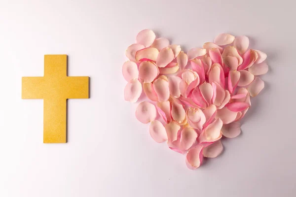 Overhead of yellow christian cross and heart shape of pink rose petals on white background. Love, faith and religion concept.