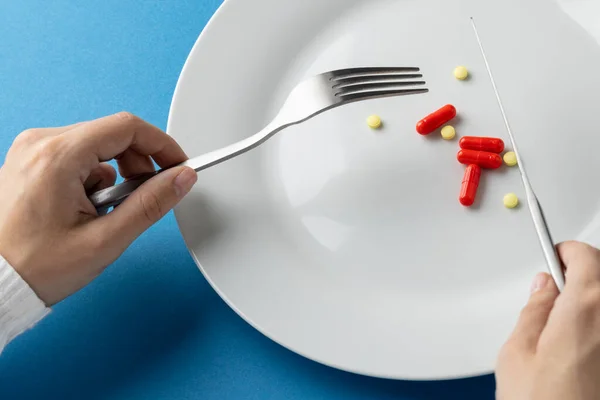 Composition of hands holding fork and knife over plate with pills, on blue background. Medicine, medical services, healthcare and health awareness concept.