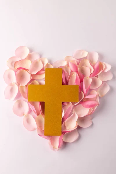 Vertical overhead of yellow christian cross on heart of pink rose petals, on white with copy space. Love, faith and religion concept.