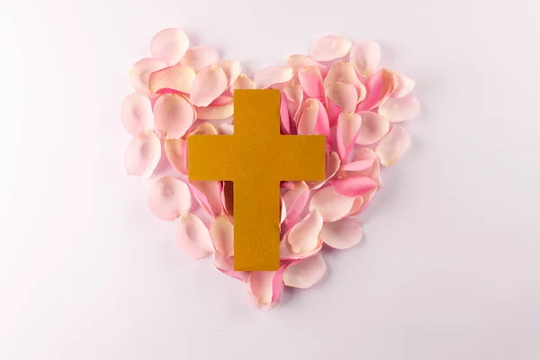 Overhead of yellow christian cross on heart shape of pink rose petals, on white background. Love, faith and religion concept.