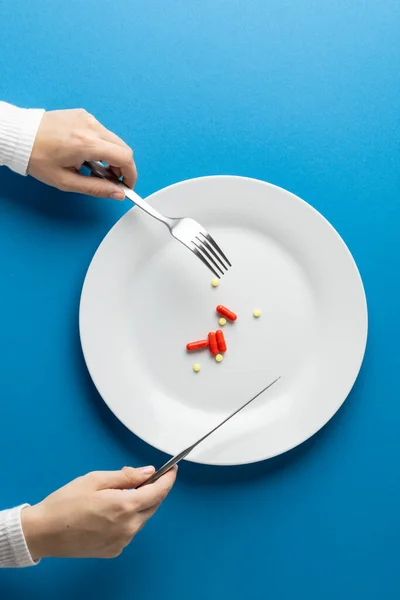 Vertical of hands holding fork and knife over plate with pills, on blue background with copy space. Medicine, medical services, healthcare and health awareness concept.