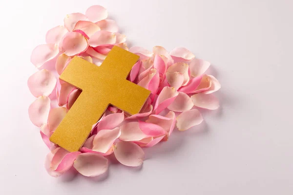 Overhead of yellow christian cross on heart shape of pink rose petals, on white with copy space. Love, faith and religion concept.