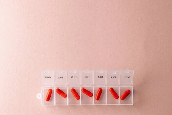 Composition of red pills in open weekly pill box on pink background with copy space. Medicine, medical services, healthcare and health awareness concept.