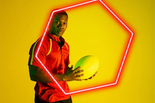 黄色の背景にボールを保持自信を持ってアフリカ系アメリカ人の若いラグビー選手のヘキサゴンネオン デジタル複合体 スポーツ ラグビー 競争力のあるスポーツ スキル コピースペース 肖像画 — ストック写真