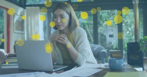 自宅のラップトップでビデオ通話をしているアジアの女性に浮かんでいる黄色の斑点のアニメーション ビジネス技術の概念 — ストック動画