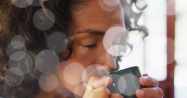 Animation of white spots of light over biracial woman drinking tea. Lifestyle, wellbeing and leisure time concept digitally generated video.