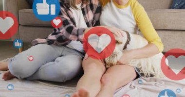 Animation of hearts and like icons over caucasian female couple sitting on sofa with their pet dog. Valentines, love and celebration concept digitally generated video.