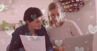Animation of hearts over caucasian female couple sitting at table and using laptop. Valentines, love and celebration concept digitally generated video.