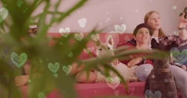 Animation of hearts over caucasian female couple sitting on sofa with their pet dogs. Valentines, love and celebration concept digitally generated video.